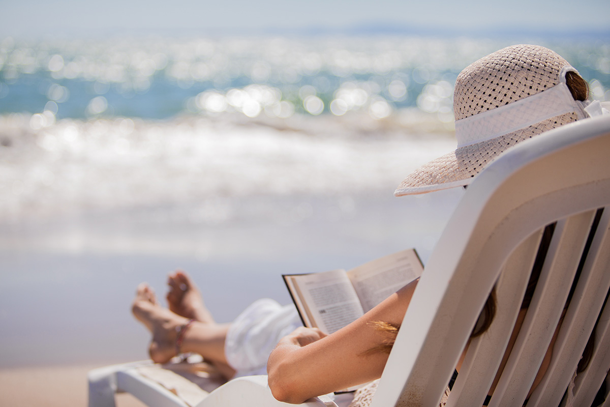 Person wearing a sunhat sits on a lounge chair at the beach, reading a book with the ocean and waves in the background.