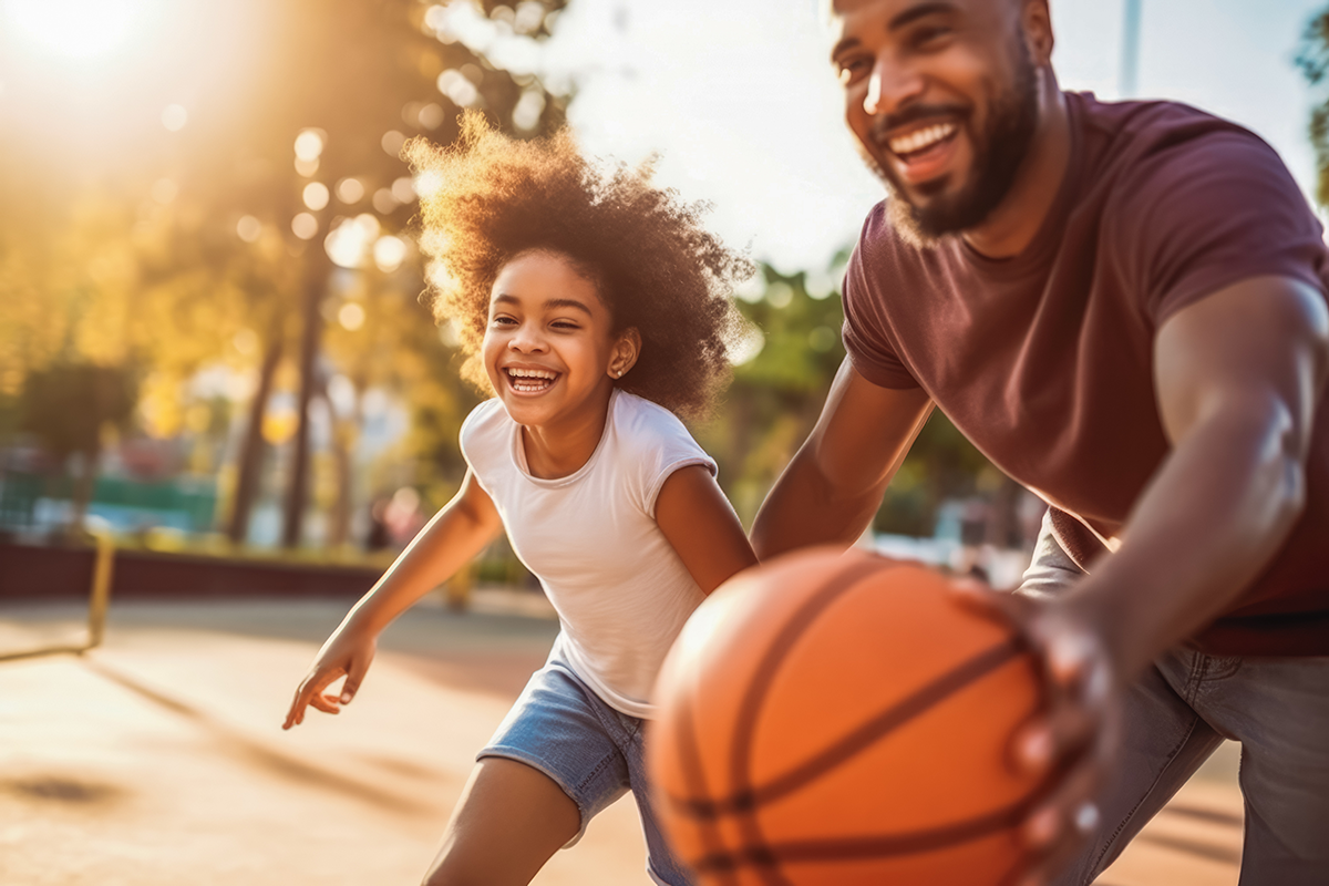 Smiling father and daughter play basketball on a sunny outdoor court, sharing a joyful moment as he dribbles the orange ball.