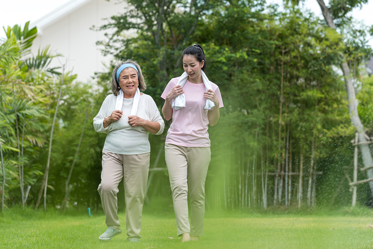 An older woman and younger woman walk side by side outdoors, smiling gently during exercise, surrounded by lush trees and garden greenery.